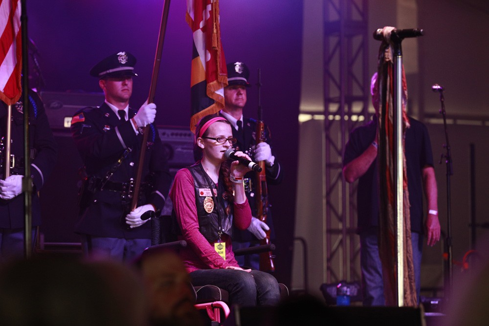 woman in wheelchair with mic on stage flanked by color guard